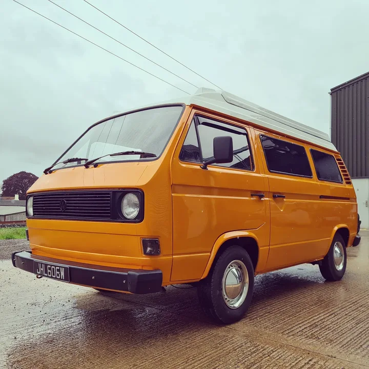 Front three-quarter view of a Volkswagen T25 camper with round headlights outside the workshop.