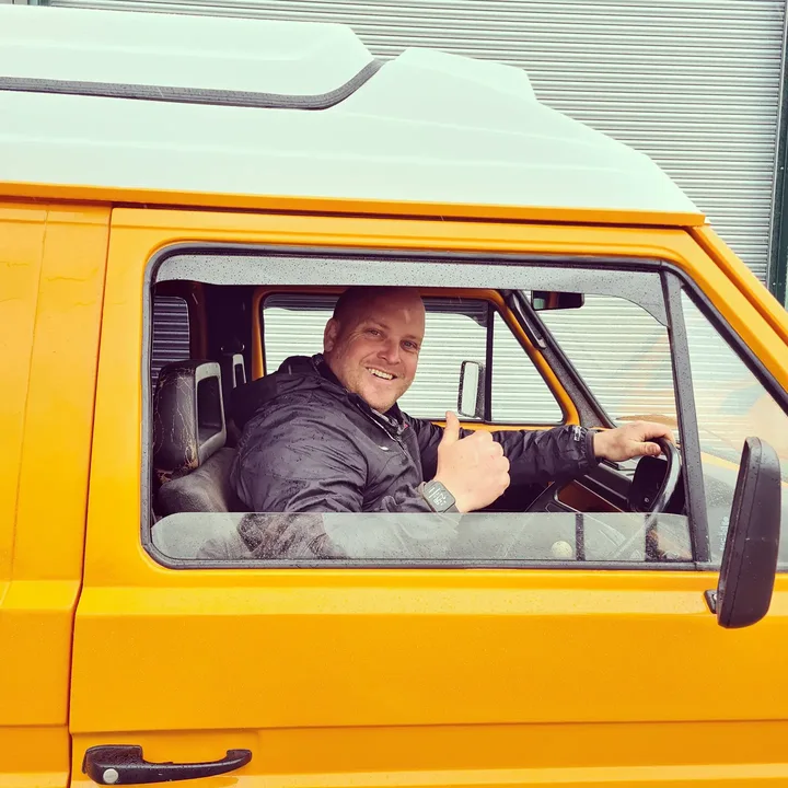 A smiling man giving a thumbs-up from the open window of a Volkswagen T25 camper.