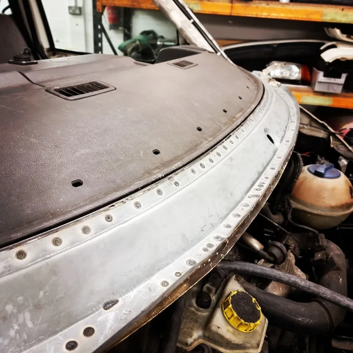 Exposed lower windscreen surround of a Volkswagen T4 Transporter with a sanded metal strip and mounting holes above the engine bay.