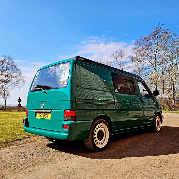 Rear three-quarter view of a Volkswagen T4 Transporter outside the workshop with sunlight reflecting along the side panels.