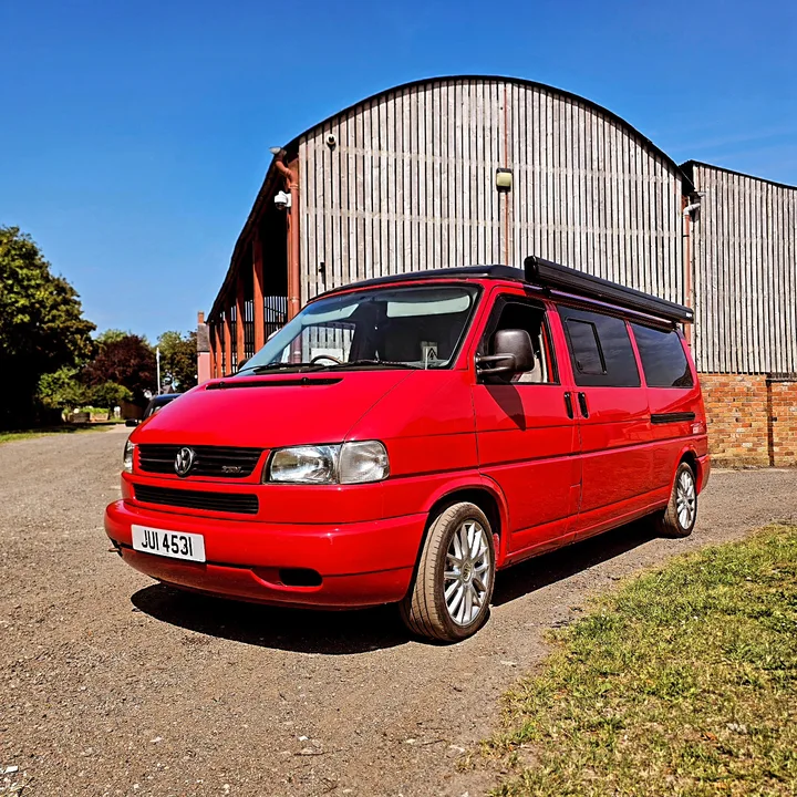 Front three-quarter view of a Volkswagen T4 camper with alloy wheels and a roof awning beside the workshop.