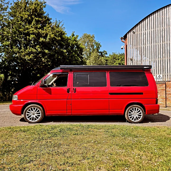 Red Volkswagen T4 Transporter with dark side windows and silver wheels parked beside a corrugated workshop.