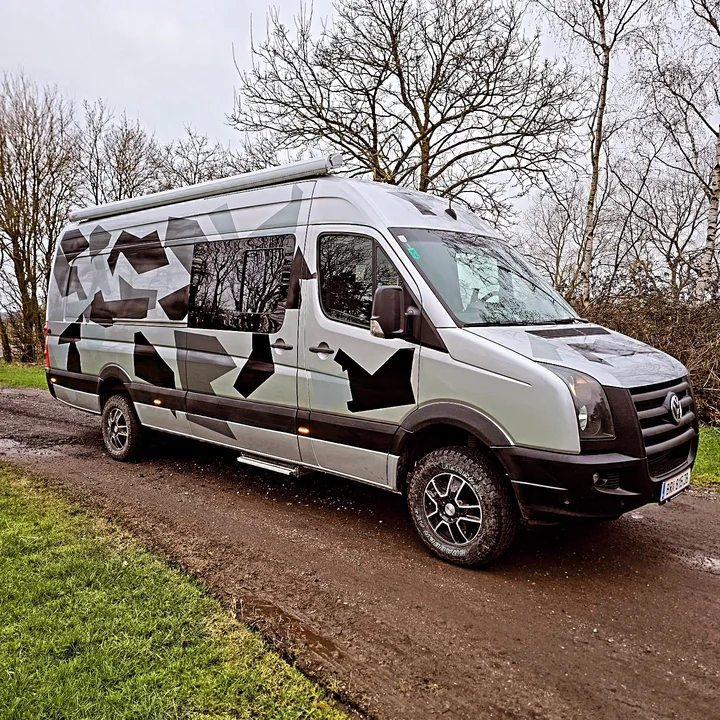 Silver Volkswagen Crafter with angular grey and black graphics parked beside trees.