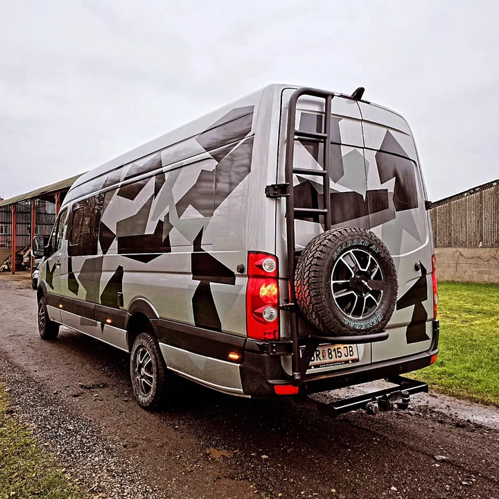Rear three-quarter view of a grey patterned Crafter beside the workshop with ladder, spare wheel and tow bar.