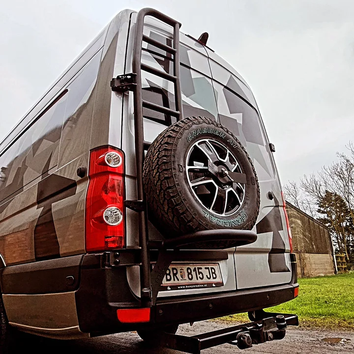Low rear view of the patterned Crafter with a black ladder, external spare wheel and tow bar.