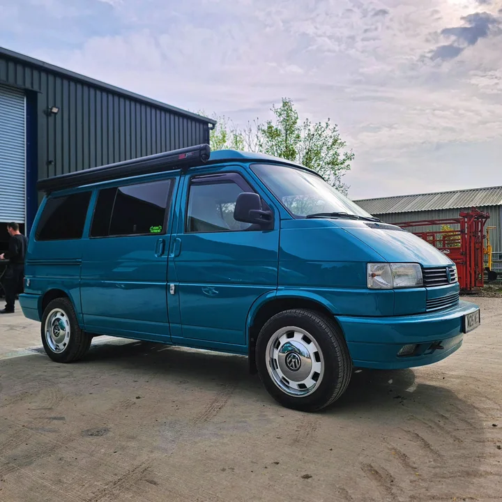 Blue T4 Transporter viewed from the front and side beside a green corrugated workshop building.
