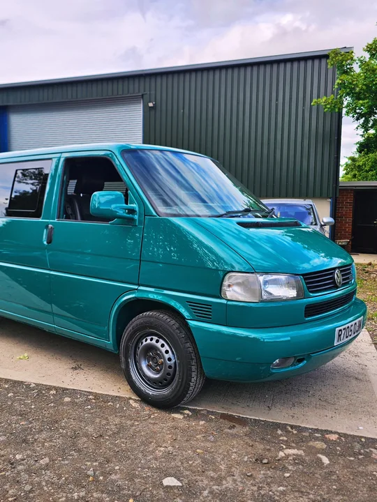 Front three-quarter view of a teal T4 Transporter with dark windows beside the workshop.