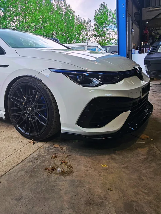 Close front three-quarter view of a white Mk8 Golf at the workshop entrance, showing its black wheel and front bumper.