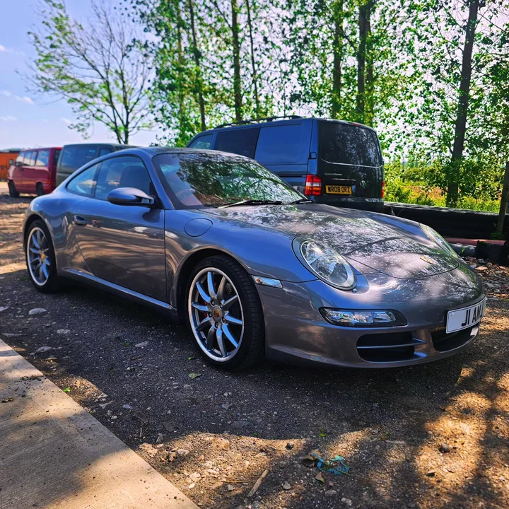 Grey Porsche 911 Carrera S coupe with silver wheels parked outside beside trees and vans.