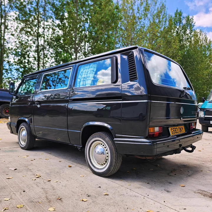 Black T25 camper photographed from the rear and side with silver wheel covers and trees behind it.