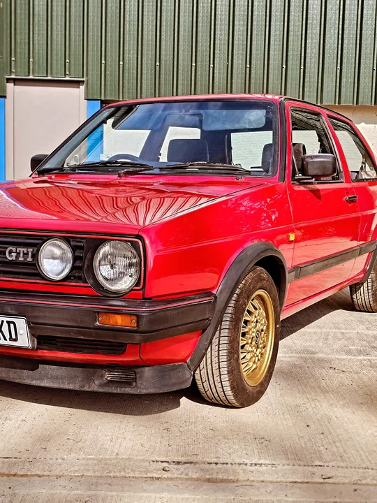 Close view of a red Mk2 Golf GTI showing its twin round headlights, GTI grille badge and gold mesh front wheel.