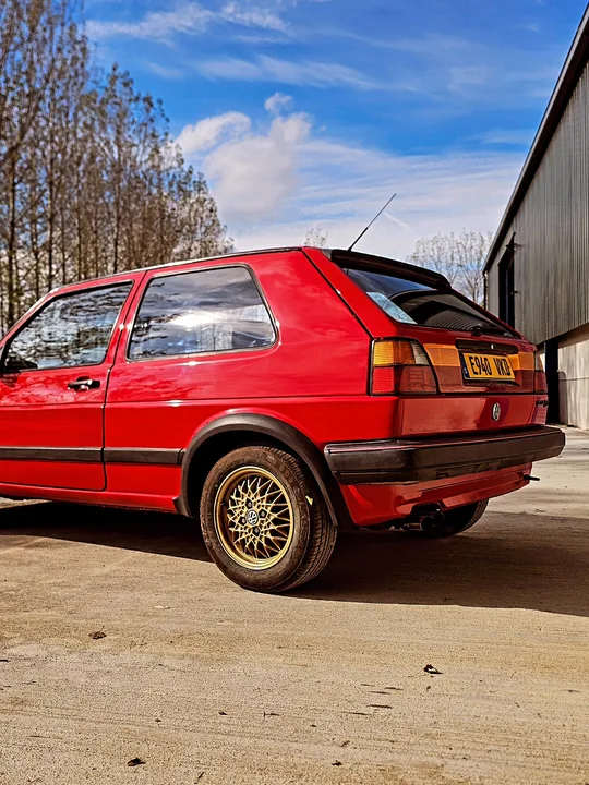 Low rear three-quarter view of a red Mk2 Golf GTI with gold mesh wheels and a rear spoiler beside the workshop.