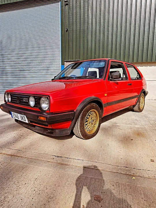 Full front three-quarter view of a red Mk2 Golf GTI with gold mesh wheels in front of a workshop roller door.