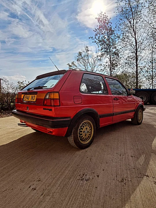 Rear three-quarter view of a red Mk2 Golf GTI with gold mesh wheels and a rear spoiler in the workshop yard.